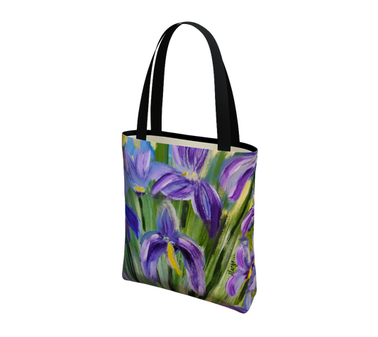 A canvas tote bag featuring a painted floral design of purple irises on a green background, with black shoulder straps, shown on a plain white background.