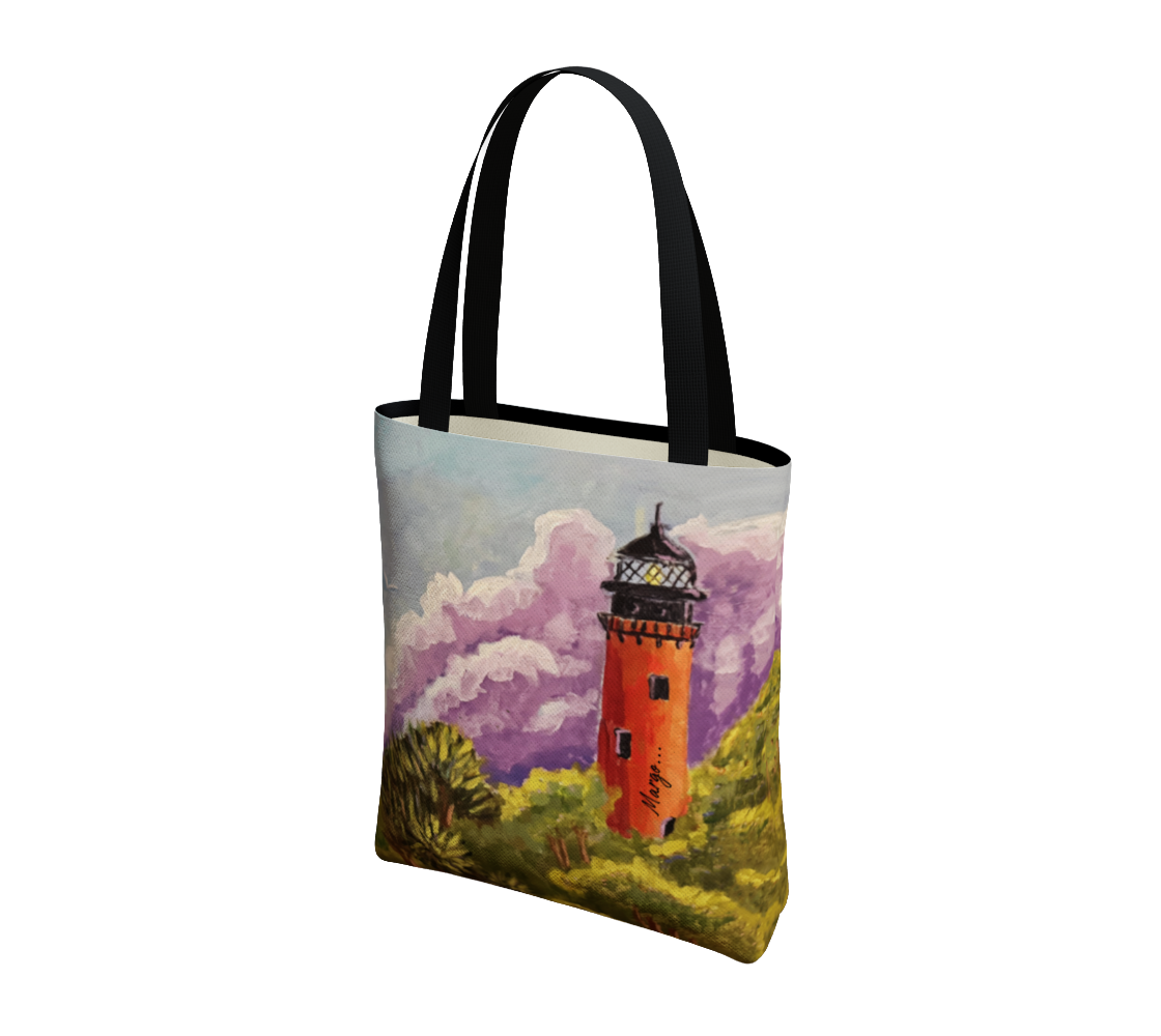 A canvas tote bag with black handles, featuring a printed artistic illustration of an orange lighthouse surrounded by green grass and purple clouds on a white background.