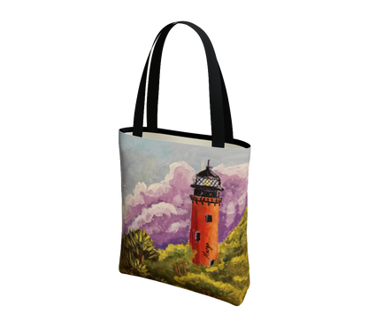 A canvas tote bag with black handles, featuring a printed artistic illustration of an orange lighthouse surrounded by green grass and purple clouds on a white background.