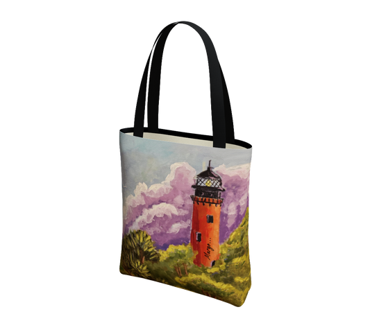 A canvas tote bag with black handles, featuring a printed artistic illustration of an orange lighthouse surrounded by green grass and purple clouds on a white background.