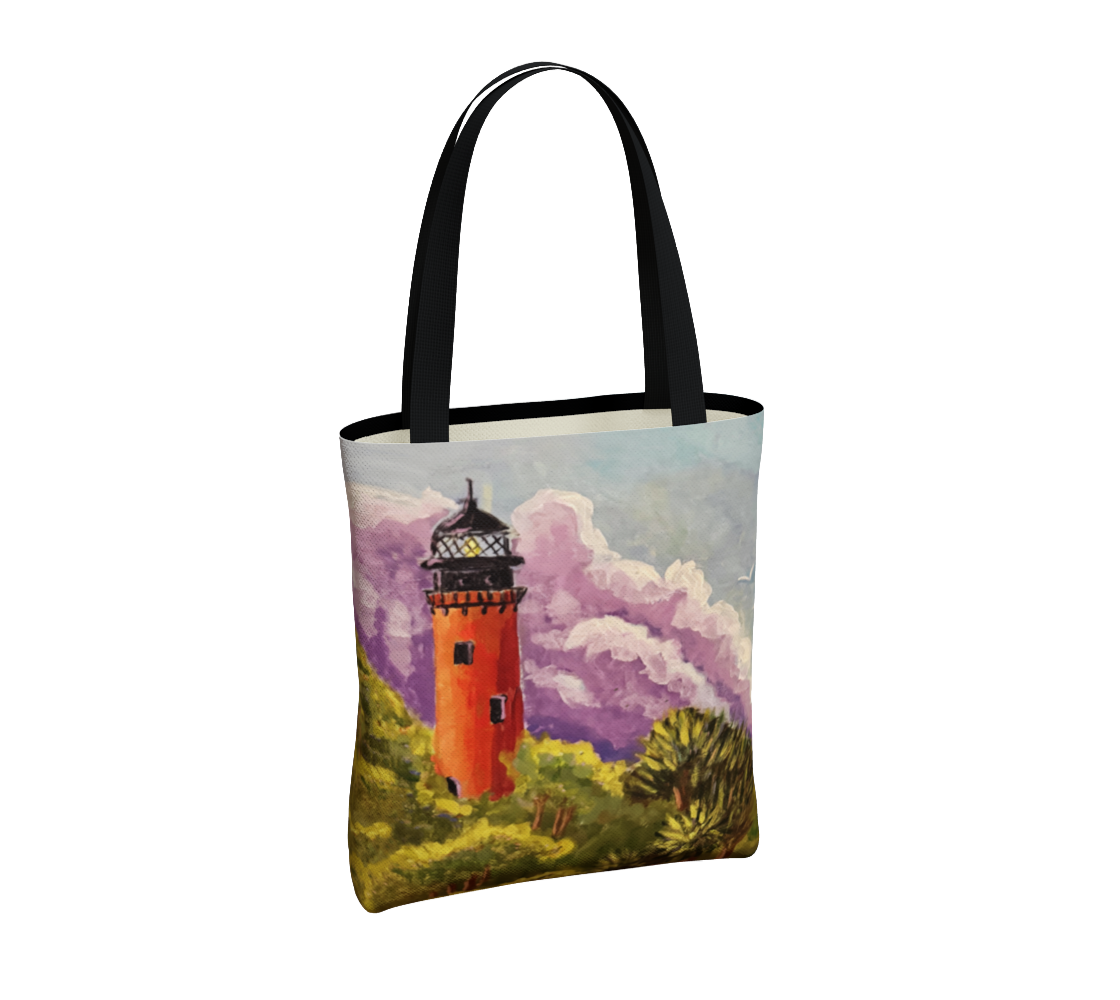 A canvas tote bag with black handles, featuring a printed illustration of an orange lighthouse surrounded by green foliage and purple clouds on a white background.