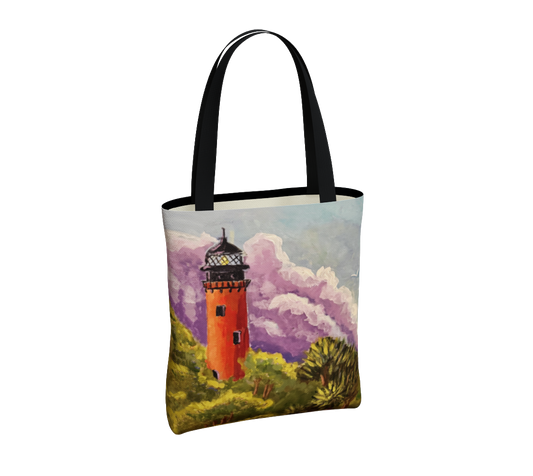 A canvas tote bag with black handles, featuring a printed illustration of an orange lighthouse surrounded by green foliage and purple clouds on a white background.
